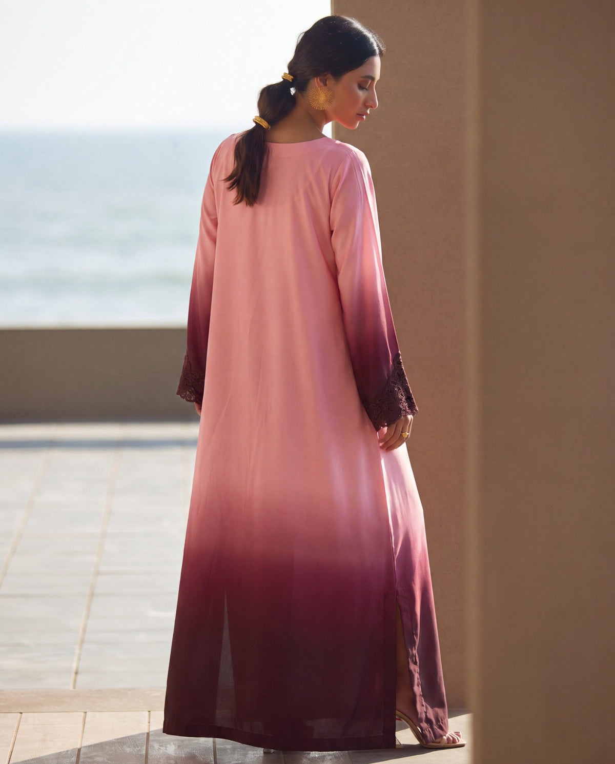 A woman wearing a long pink abaya with a dark burgundy ombre gradient at the hem and cuffs, standing outdoors near a wall with a blurred sea background.